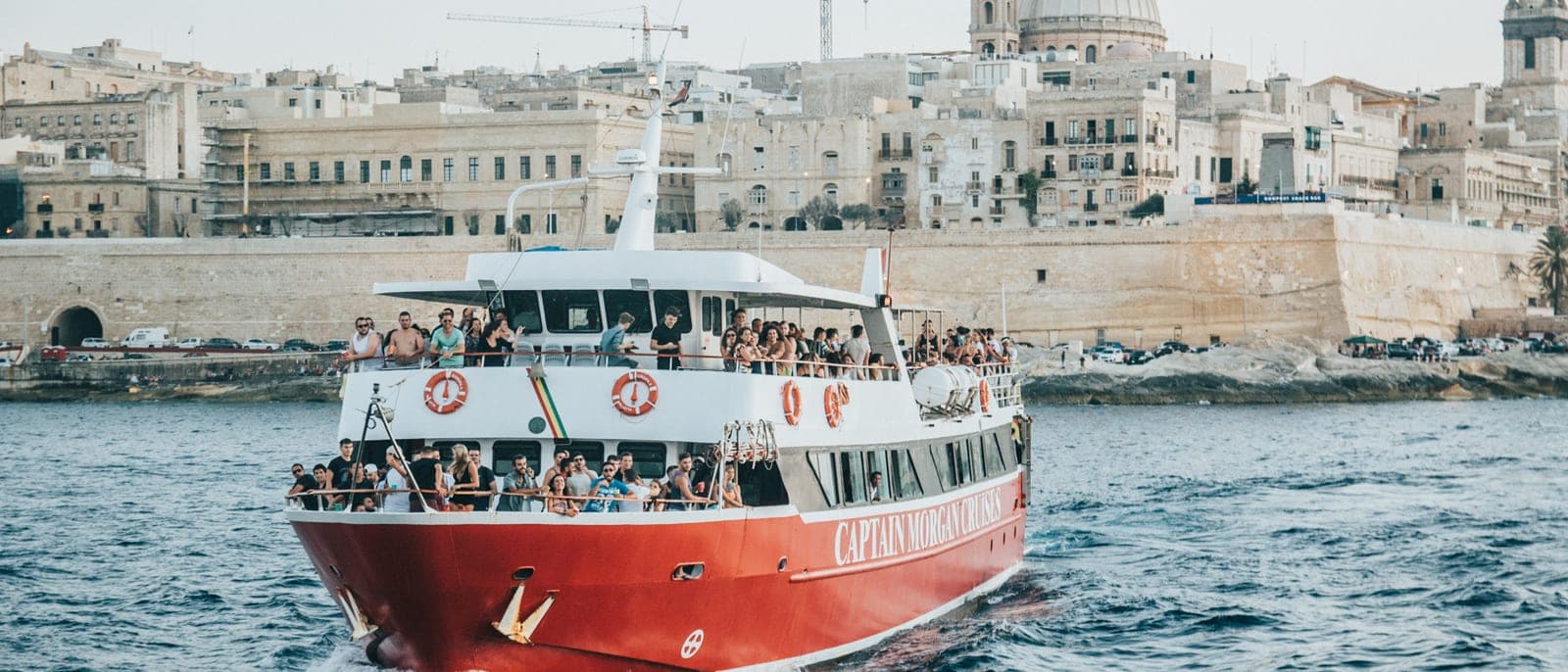 Un barco de excursión lleno de gente llegando al puerto con las murallas y la cúpula de La Valeta detrás, a media tarde