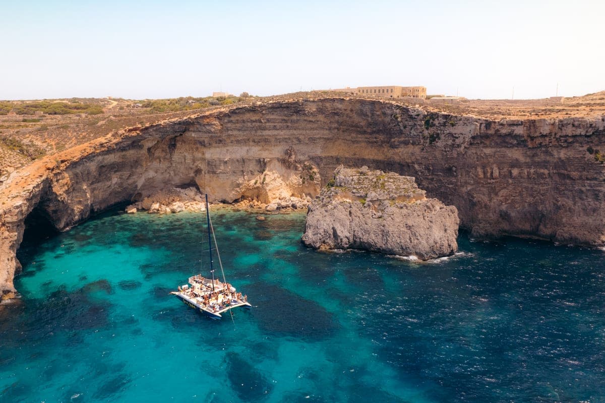 Una cala de Comino desde el aire con un solo catamarán fondeado en el agua turquesa, acantilados bajos alrededor y sin gente