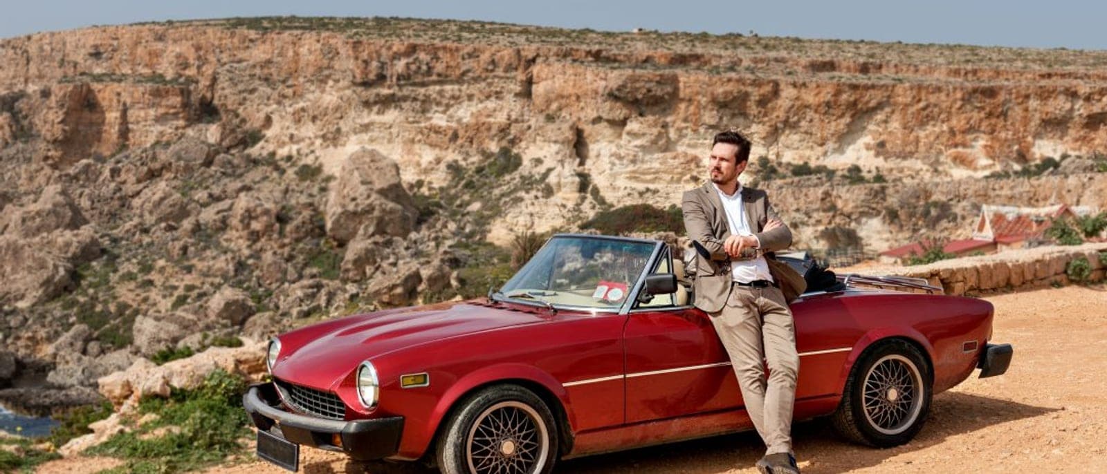Un descapotable rojo aparcado en un mirador sobre una bahía de acantilados de Malta, con un hombre apoyado en la puerta