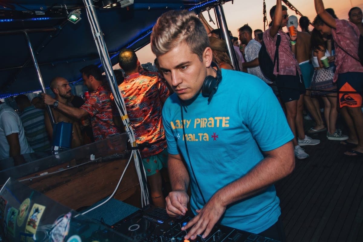 El DJ pinchando en la cubierta principal de la Lazy Pirate al atardecer, con la gente bailando detrás