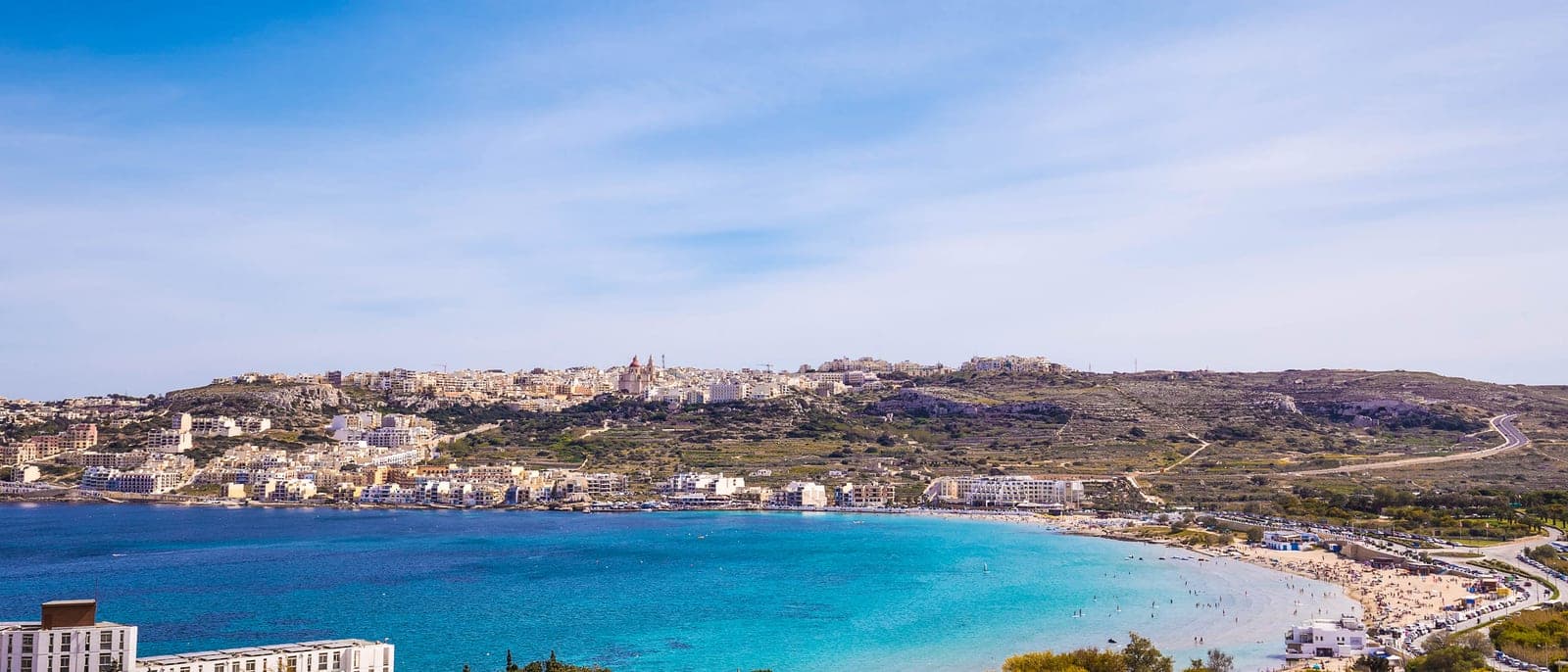 La bahía de Mellieħa desde lo alto de la carretera que baja del pueblo: la playa larga de arena de Għadira, el agua turquesa y poco profunda y, al fondo, la colina con la Torre Roja