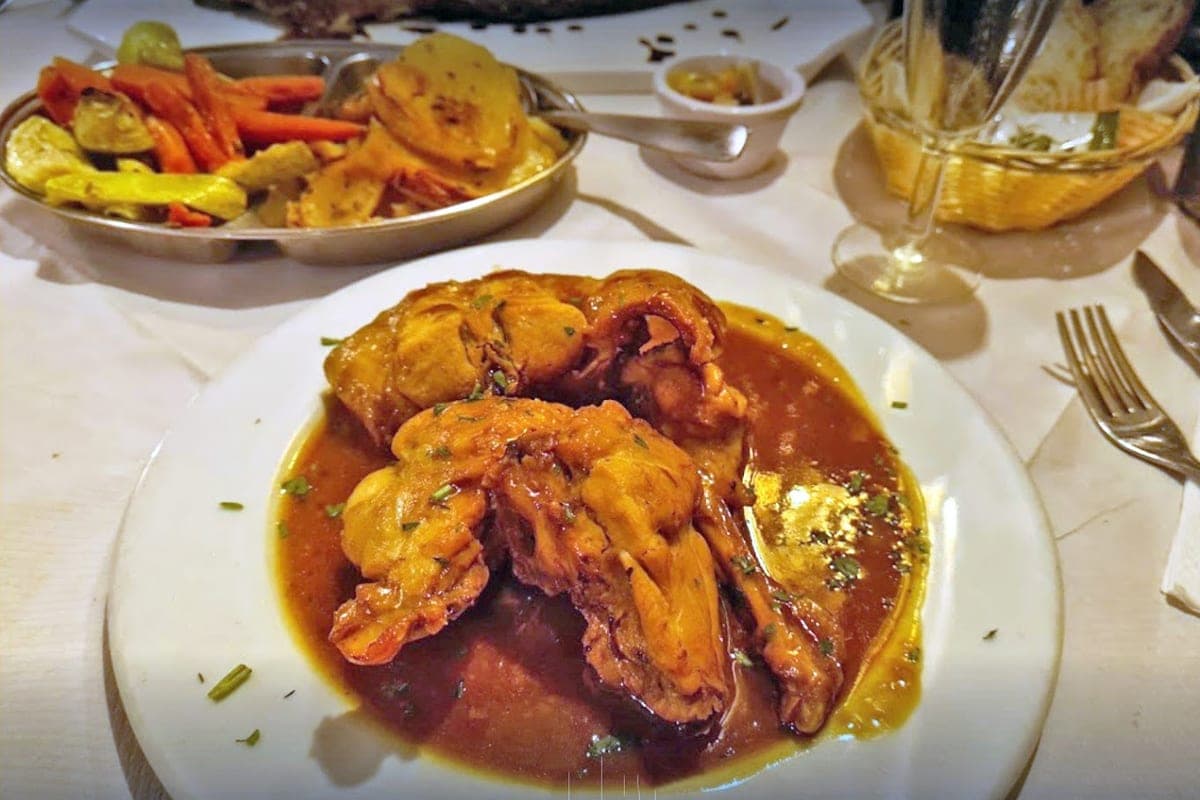 Un plato de conejo guisado maltés, el stuffat tal-fenek, con su salsa oscura, en una mesa de restaurante con patatas y verdura al lado