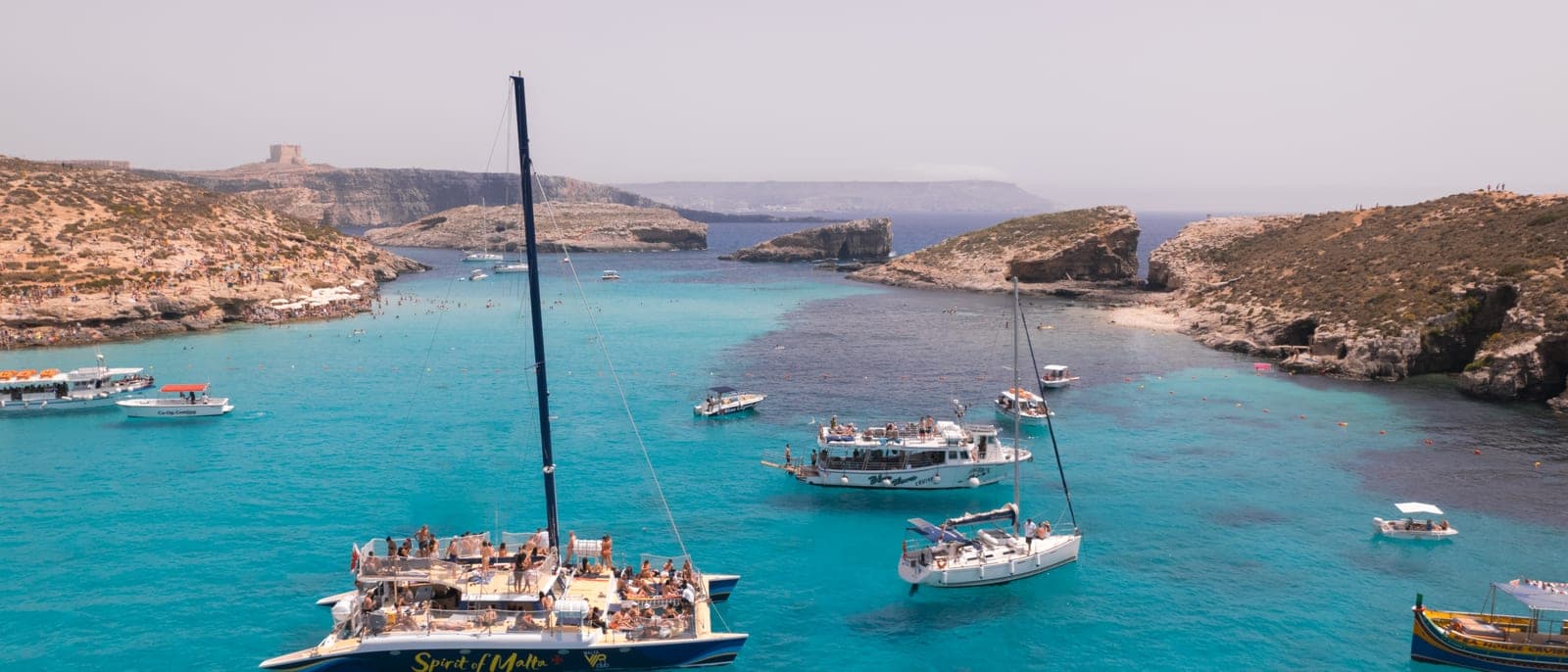 La Blue Lagoon de Comino desde el aire: el agua turquesa entre Comino y Cominotto con varios barcos y catamaranes fondeados, y la torre de vigilancia en la colina