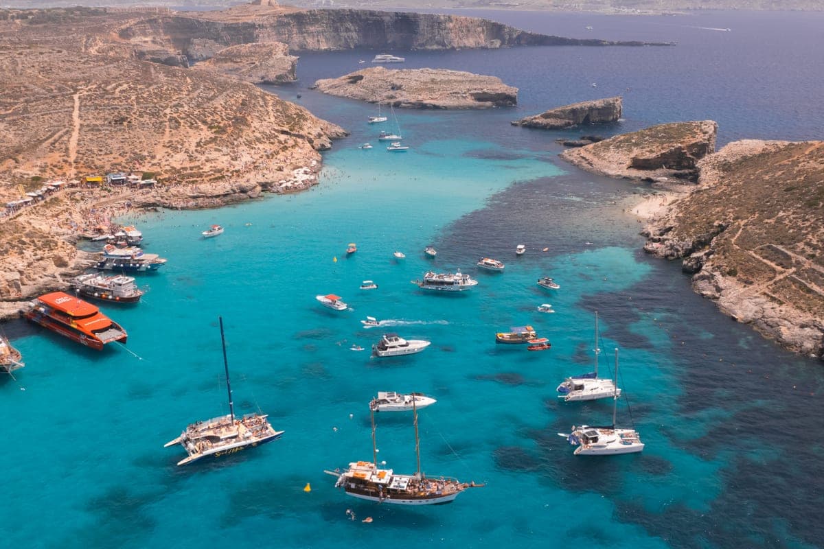 La Blue Lagoon desde el aire a media tarde: decenas de barcos y catamaranes fondeados en el paso entre Comino y Cominotto y la roca llena de gente