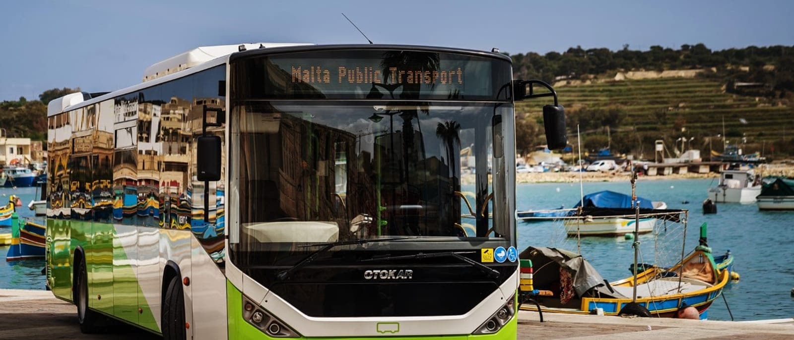 Un autobús verde y blanco de Malta Public Transport parado junto al puerto de un pueblo de pescadores, con las barcas de colores detrás