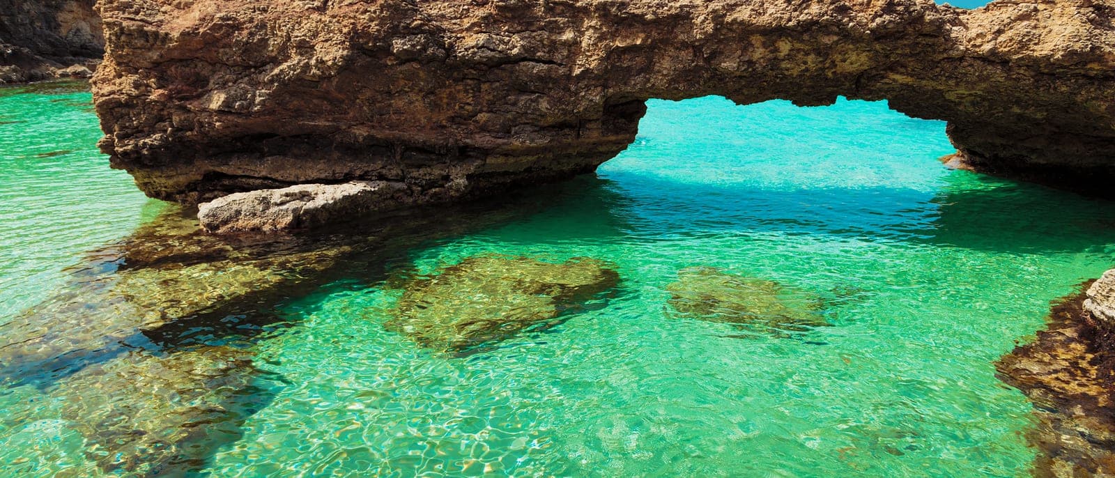 Un arco de roca sobre el agua turquesa y transparente de una cala de Comino, sin nadie, con el fondo de arena y piedra visible