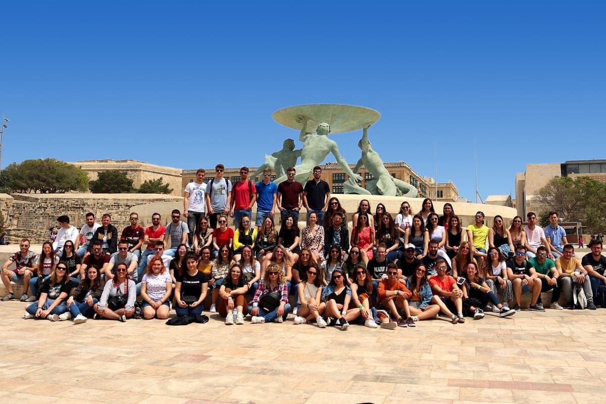 Un grupo grande de estudiantes jóvenes sentados y de pie delante de la Fuente de los Tritones, a la entrada de La Valeta