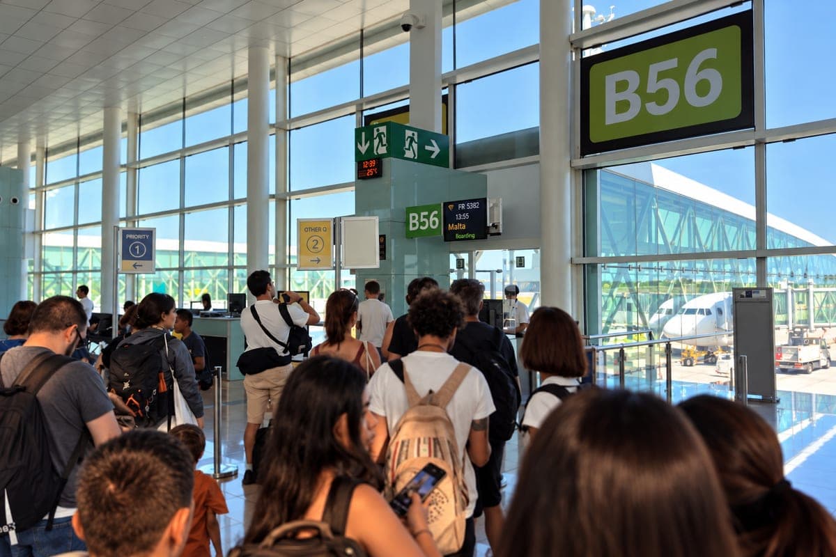 La cola de embarque de un vuelo a Malta en la puerta B56 de un aeropuerto español, con los pasajeros de espaldas con sus mochilas, el cartel del vuelo y el avión al otro lado del cristal