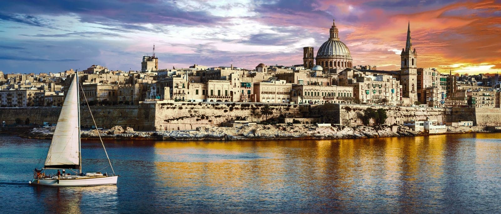 La Valeta vista desde Sliema al atardecer, con las cúpulas y las murallas doradas por el sol y un velero cruzando el puerto de Marsamxett