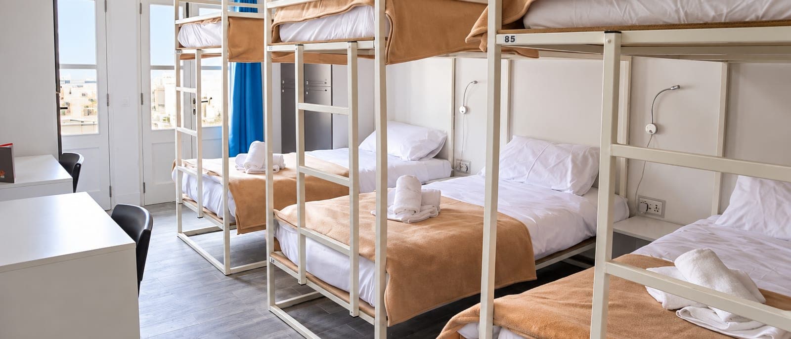 Una habitación compartida de un hostel de Sliema, limpia y luminosa, con literas de madera, taquillas y una ventana con un balcón maltés de colores enfrente