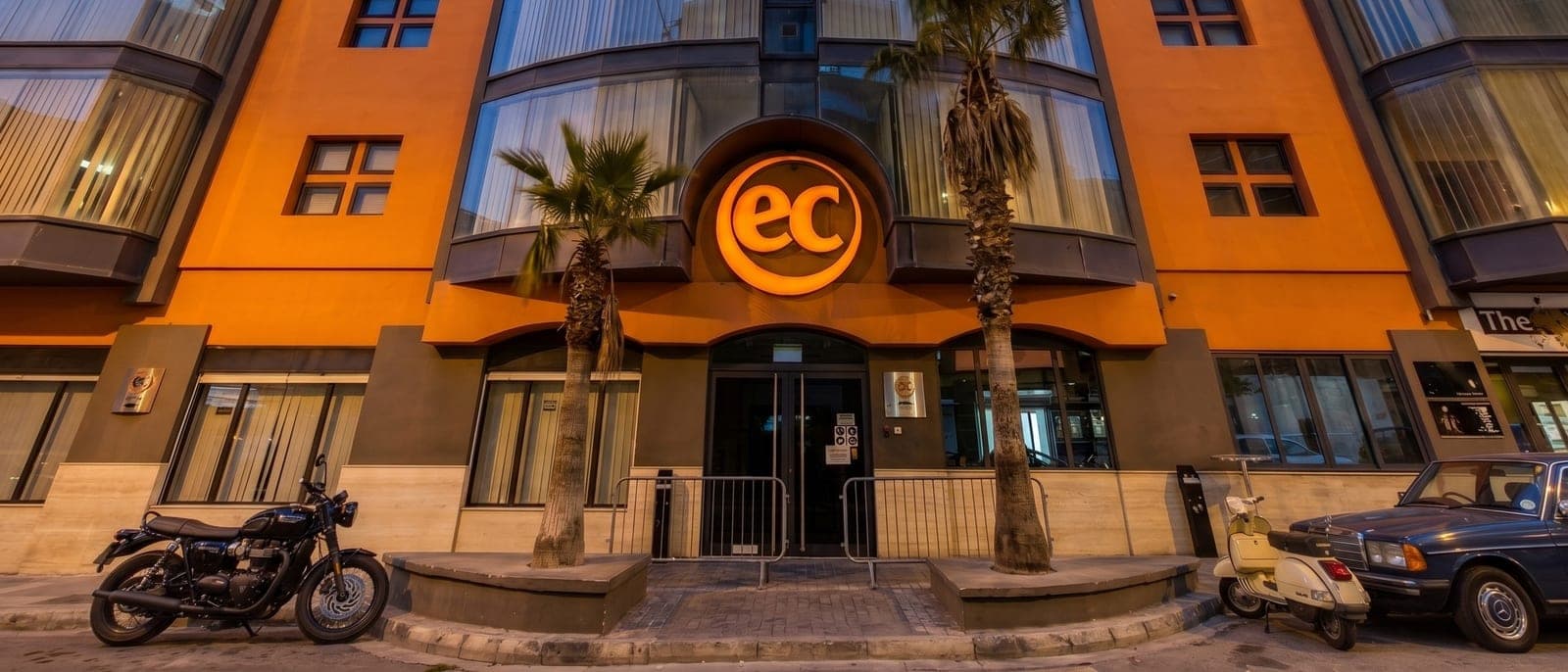 La fachada de EC Malta en Marguerite Mangion Street, St Julian's, al anochecer: el rótulo naranja de EC sobre la entrada, con las puertas cerradas y vallas delante