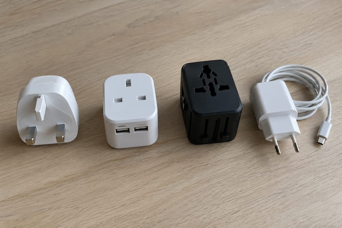 Sobre una mesa, tres adaptadores de viaje distintos: uno sencillo blanco, uno con tomas USB y uno universal, junto a un cargador español