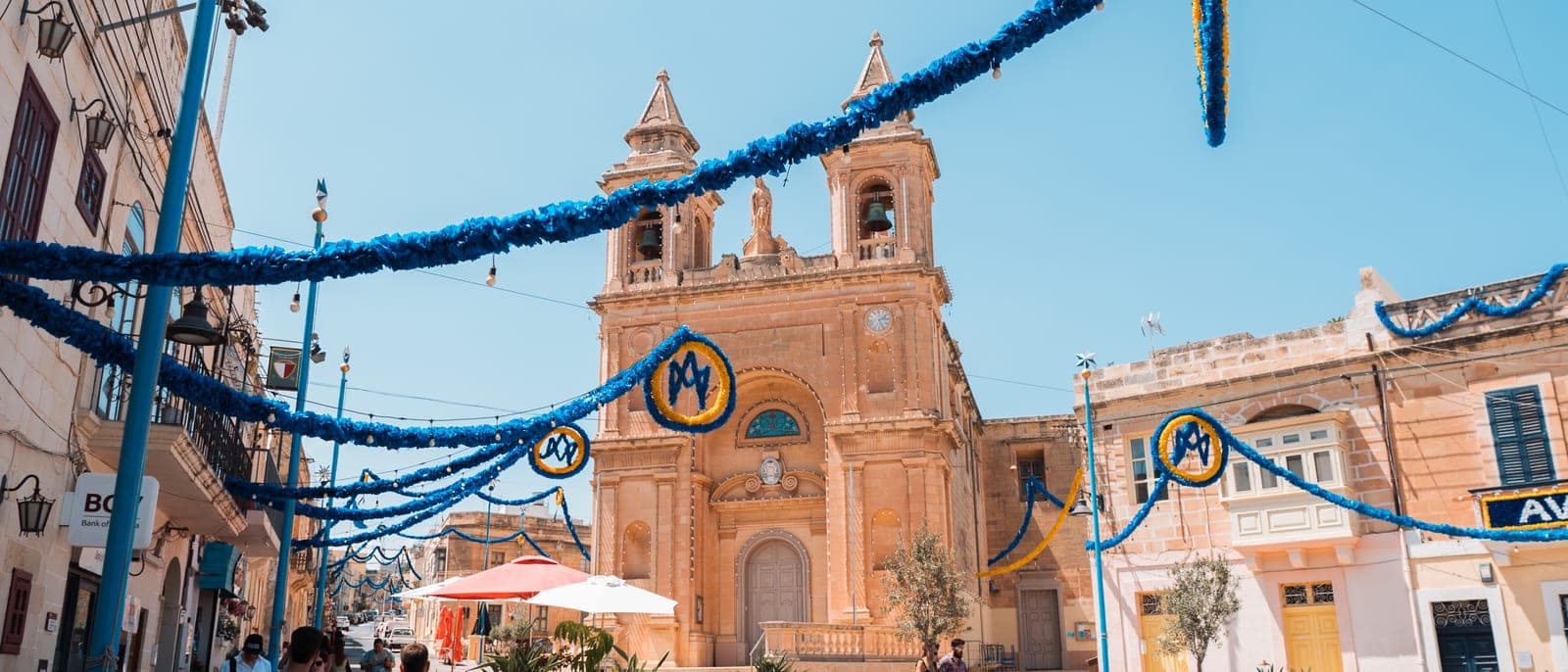 La plaza de la iglesia de Marsaxlokk con los adornos de festa colgados: guirnaldas azules entre los postes, escudos con la M de la Virgen y la fachada de la parroquia