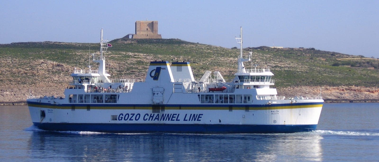 El ferry de Gozo Channel en el canal, con la torre de Santa María de Comino en la colina detrás y gente en la cubierta