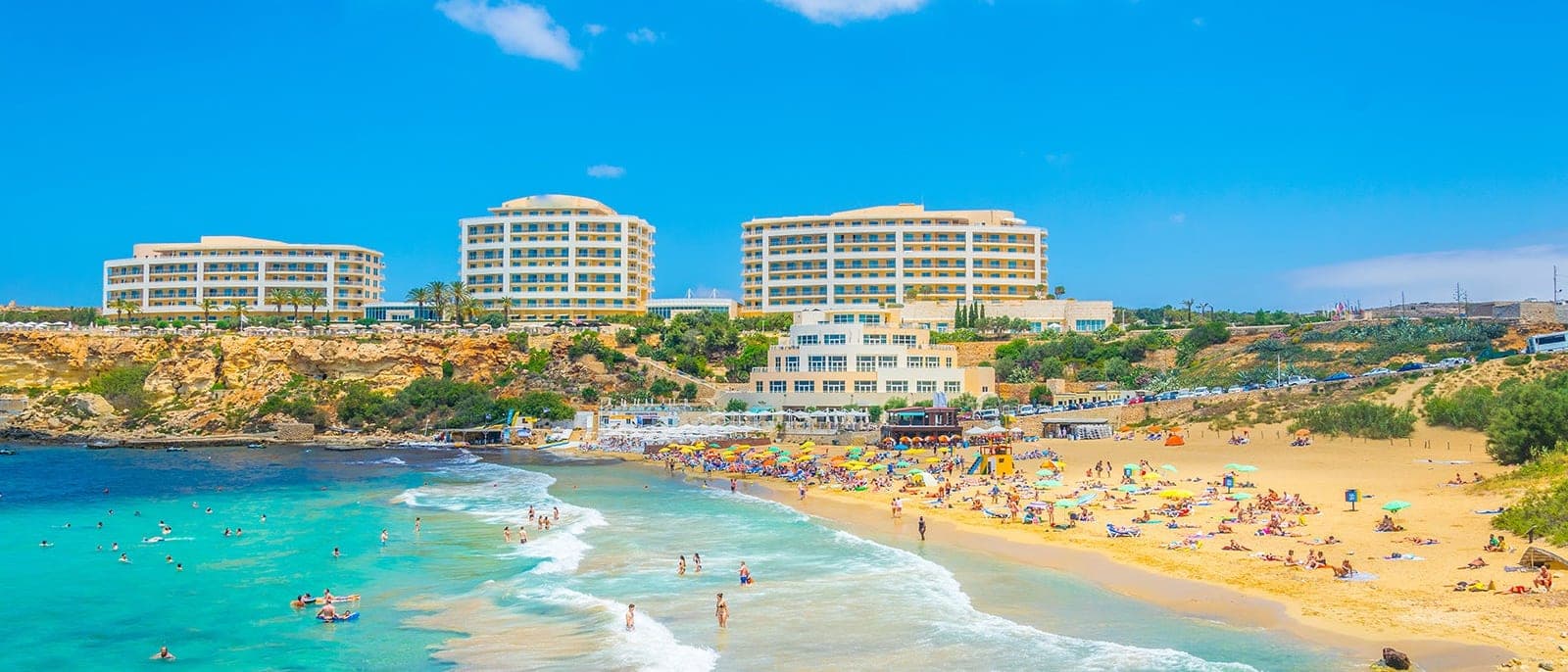 Golden Bay desde lo alto: la playa de arena dorada llena de gente y sombrillas, el mar turquesa y el hotel grande sobre el acantilado