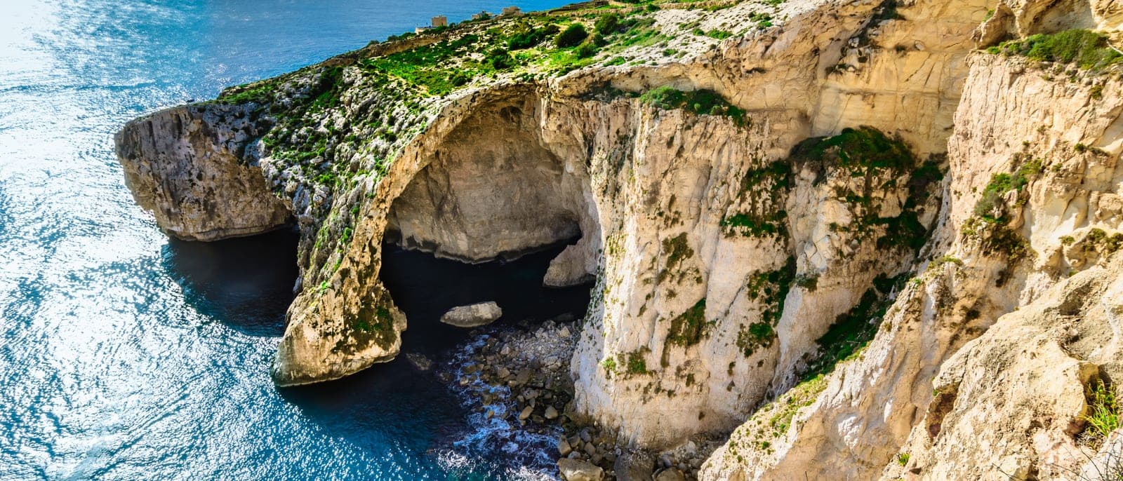 El arco de la Gruta Azul desde el mirador de la carretera: la roca blanca sobre el mar azul intenso, con las lanchas pasando por debajo y los acantilados de Wied iż-Żurrieq