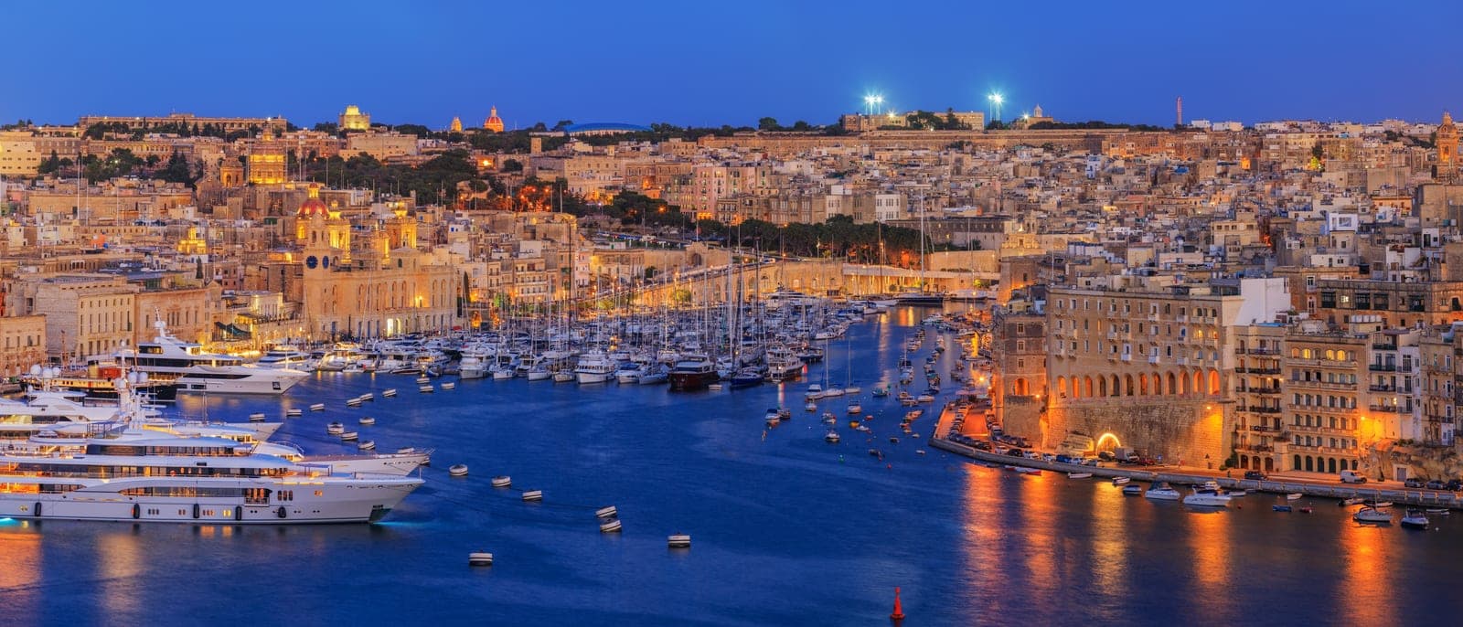 El Grand Harbour de noche desde lo alto: las Tres Ciudades iluminadas, los yates en el puerto y las murallas doradas por las farolas