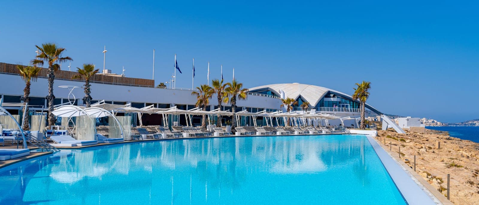 La piscina grande de un complejo de la costa de Malta al mediodía, con tumbonas y sombrillas en fila, palmeras y el mar al fondo