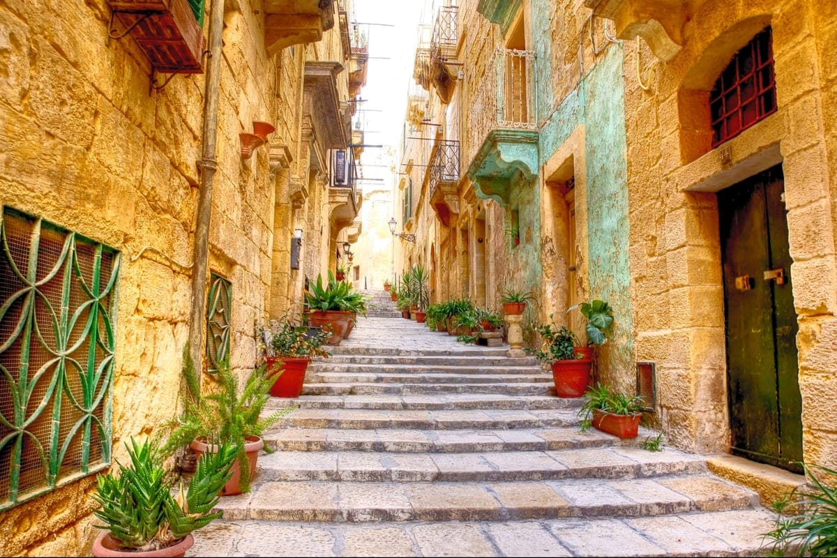 Una calle de escaleras de La Valeta bajando entre fachadas de piedra dorada, con macetas en los escalones y balcones cerrados de colores