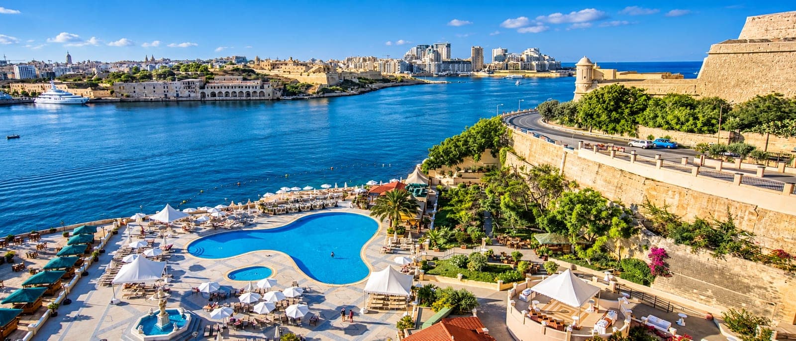 La piscina de un hotel al pie de los bastiones de La Valeta, con tumbonas y sombrillas, y enfrente el puerto de Marsamxett con Sliema al fondo