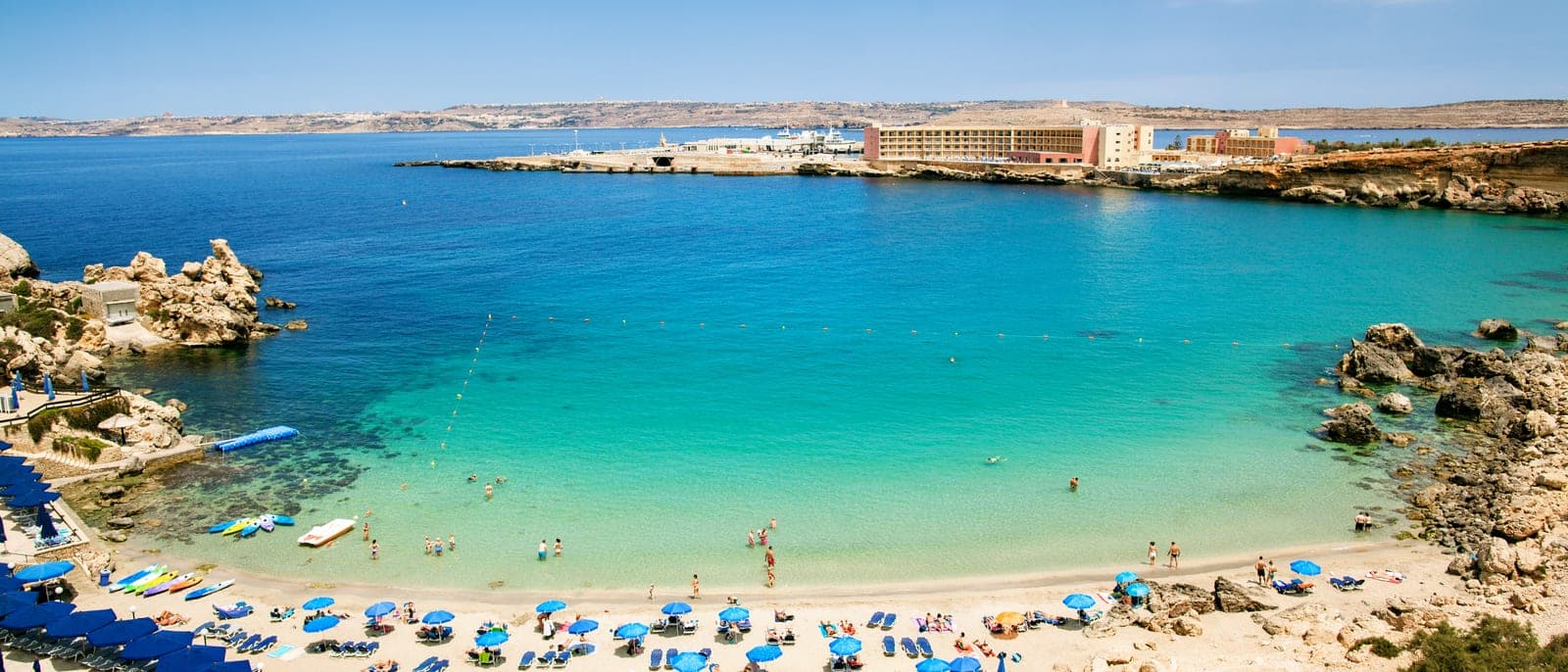 Paradise Bay desde arriba: una cala pequeña de arena con sombrillas y familias, el agua turquesa y poco profunda, y la terminal del ferry de Gozo al fondo