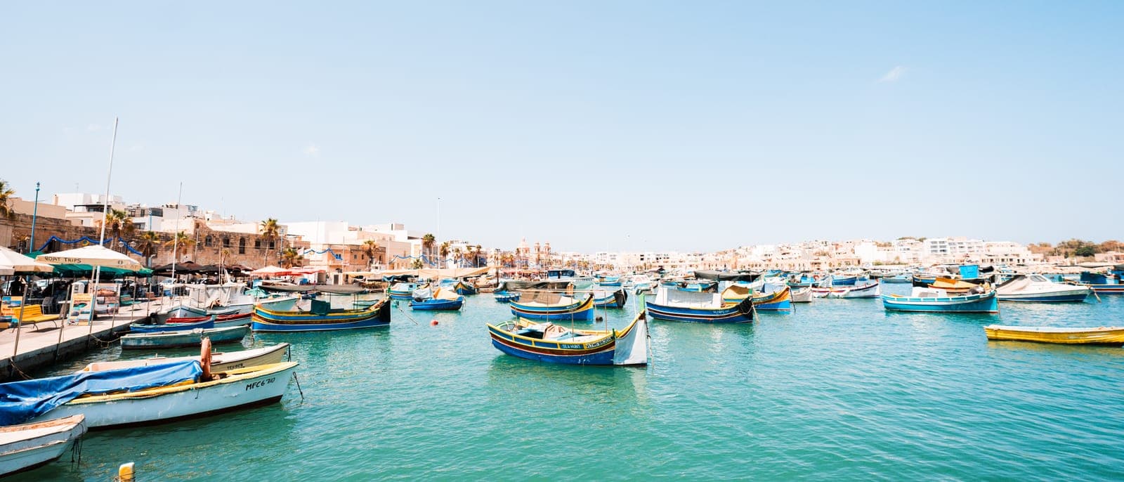 El puerto de Marsaxlokk con decenas de barcas luzzu de colores amarradas en el agua verde y las casas bajas del pueblo detrás, en un día de sol