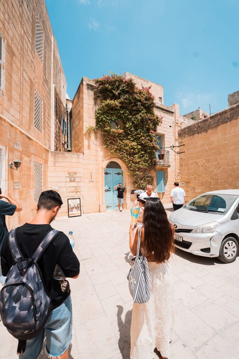 Una calle de Mdina con fachadas de piedra dorada, una enredadera sobre una puerta azul y visitantes andando hacia el fondo