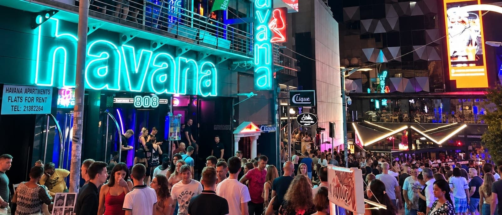 St George's Road de noche, llena de gente joven de acera a acera, con el neón turquesa del Havana y su terraza arriba, el rótulo del Clique a la derecha y las pantallas de los locales al fondo