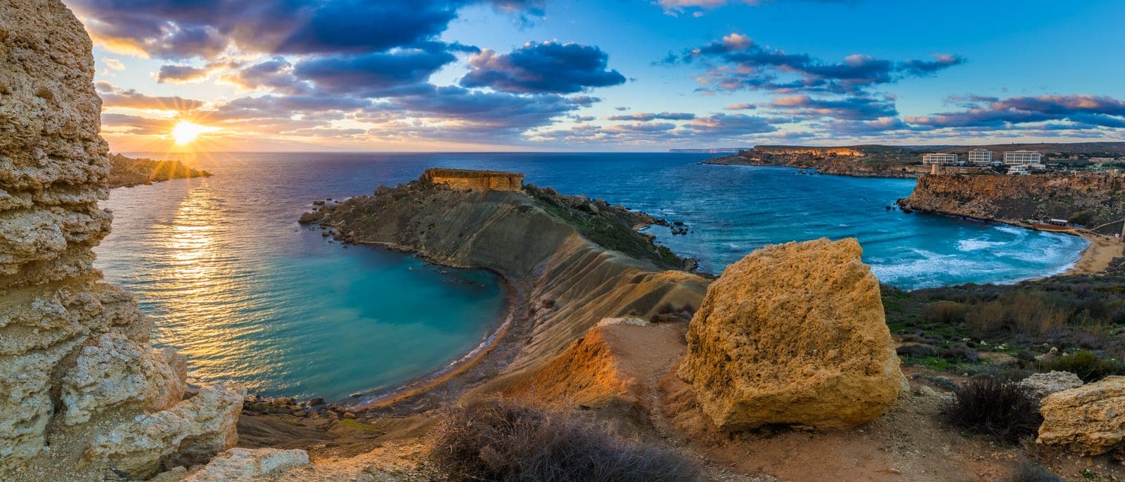 La bahía de Għajn Tuffieħa al atardecer desde lo alto de la costa: la playa de arena rojiza en media luna, el promontorio de arcilla y el sol bajando sobre el mar