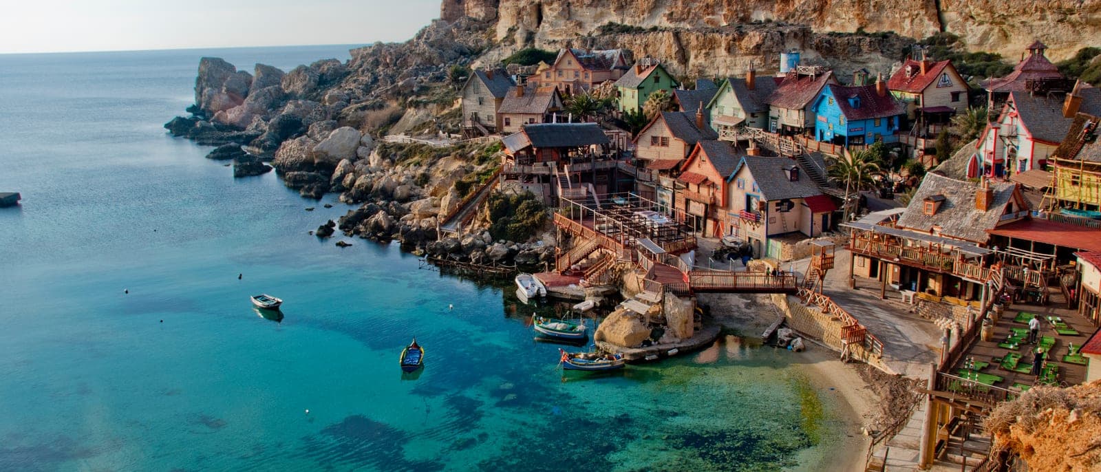Popeye Village desde el mirador de la carretera: las casas de madera de colores del decorado en la cala de Anchor Bay, el agua turquesa y el acantilado detrás