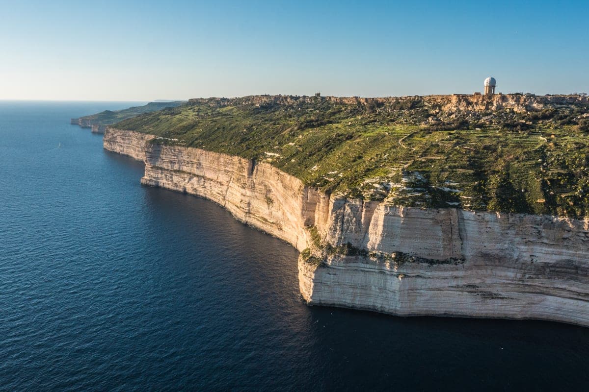 Los acantilados de Dingli desde el aire: la pared de roca blanca cayendo al mar azul oscuro, el campo verde arriba y la capilla en el borde