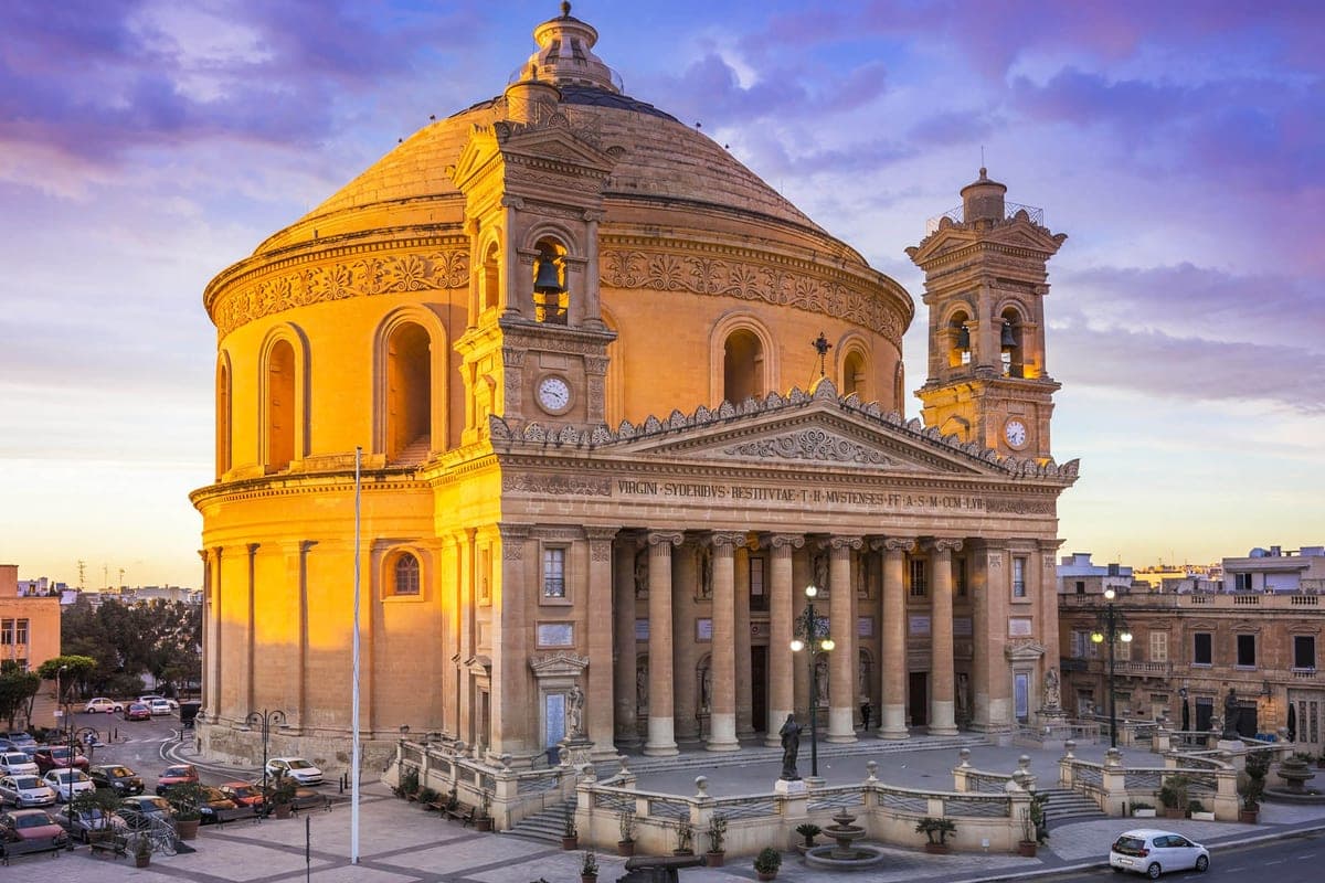 La Rotonda de Mosta, la iglesia de la gran cúpula, vista de frente desde la plaza a última hora de la tarde, con coches pequeños delante que dan la escala