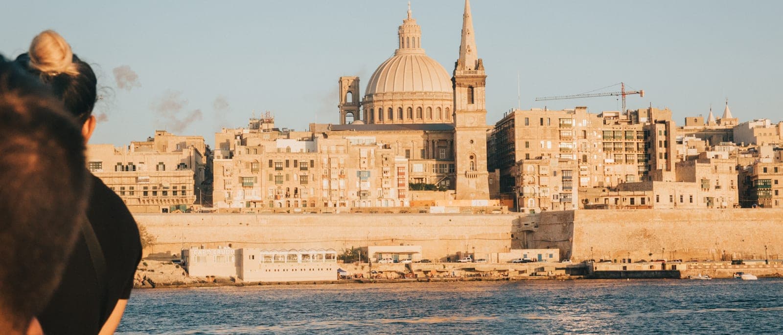La Valeta vista desde el barco que cruza desde Sliema, con un pasajero en primer plano, la cúpula del Carmen, el campanario de San Pablo y las murallas