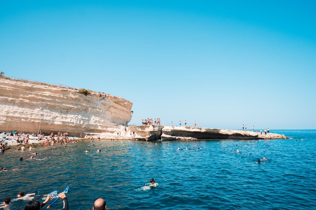 La plataforma baja de roca de St Peter's Pool con gente bañándose, el borde blanco de la bahía y el mar abierto detrás