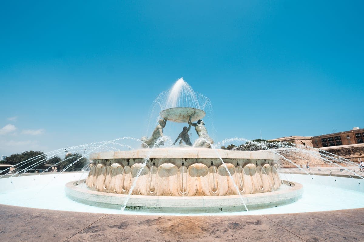 La fuente del Tritón a la entrada de La Valeta, con los chorros de agua y el cielo azul, junto a la terminal de autobuses y la parada de taxis