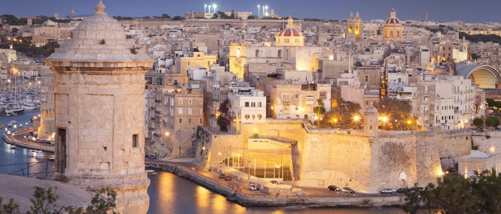 Las Tres Ciudades de noche, con una garita de piedra de la muralla en primer plano, las casas y las iglesias iluminadas y los yates en el agua