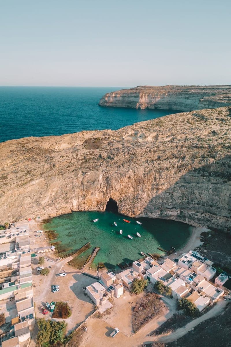 El Mar Interior de Dwejra, en Gozo, desde el aire: la laguna verde con barcas, el túnel en la roca que sale al mar, los cobertizos de los pescadores y el acantilado con el mar abierto detrás