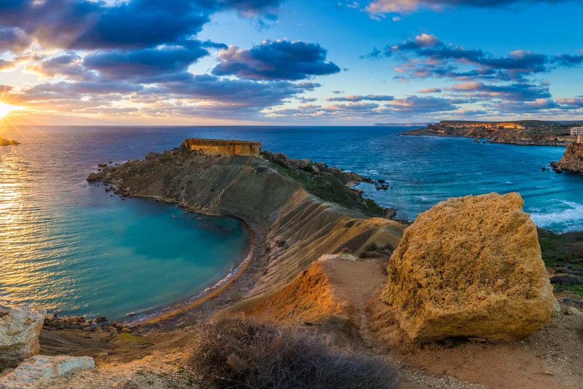 Għajn Tuffieħa al atardecer desde lo alto de la costa, con la bahía entera de arena rojiza, el promontorio y el mar