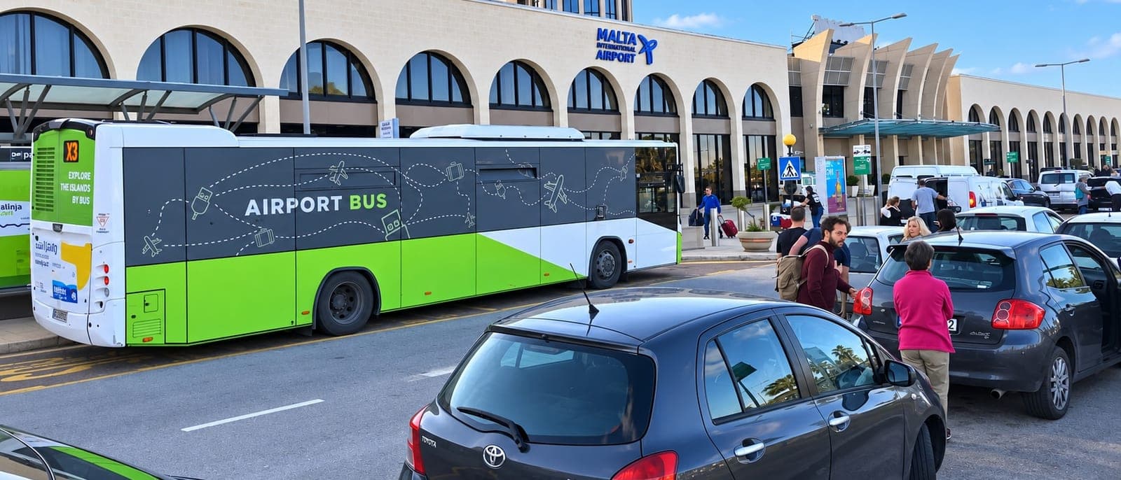La fachada de arcos de la terminal del aeropuerto de Malta con el rótulo Malta International Airport, un Airport Bus verde y blanco en la parada y viajeros con mochilas y maletas junto a los coches de la zona de recogida