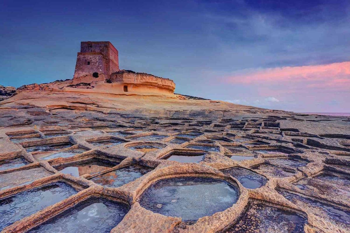 Las salinas de Xwejni junto a Marsalforn al amanecer: el tablero de piedra con los cuadrados de agua reflejando el cielo rosado y la torre de vigilancia detrás